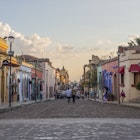 Two men stand in the middle of a cobble stone street lined with colorful buildings and street lamps some people walk on the sidewalks the sun is setting casting a golden glow in LGBTQ-friendly Oaxaca