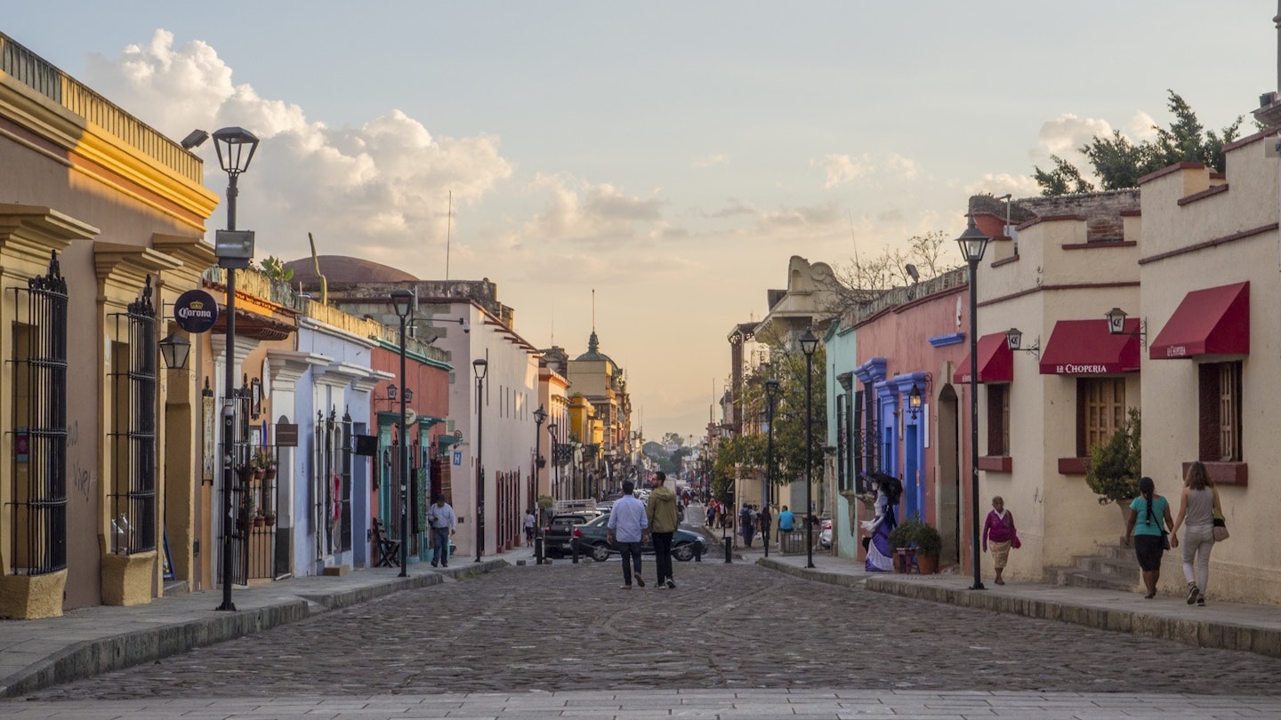 Two men stand in the middle of a cobble stone street lined with colorful buildings and street lamps some people walk on the sidewalks the sun is setting casting a golden glow in LGBTQ-friendly Oaxaca