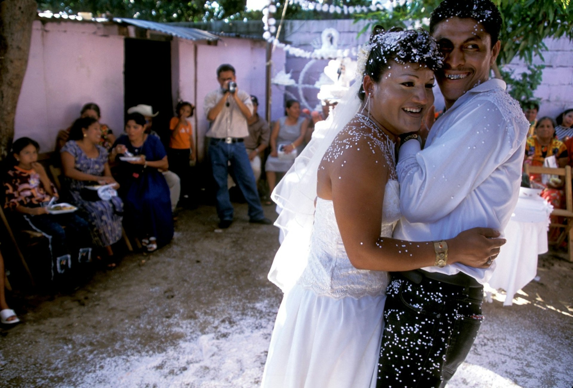 Two men, one dressed in white and the other in a dress shirt and pants hug at their wedding being photographed by people sitting on chairs in a courtyard