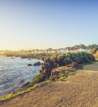 a sandy path leads past a rocky shoreline lined with wild fauna with stately homes in the background in Monterey, California