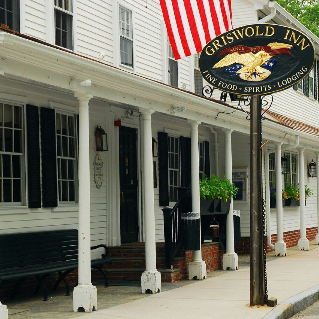 A white building with a long collonade held up by white bosts, the Griswold Inn is marked by an oval sign with an eagle on it and an American Flag flying above
