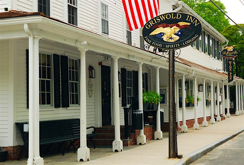 A white building with a long collonade held up by white bosts, the Griswold Inn is marked by an oval sign with an eagle on it and an American Flag flying above. Its one of the most historic taverns in the country.