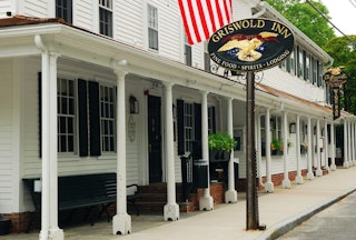 A white building with a long collonade held up by white bosts, the Griswold Inn is marked by an oval sign with an eagle on it and an American Flag flying above