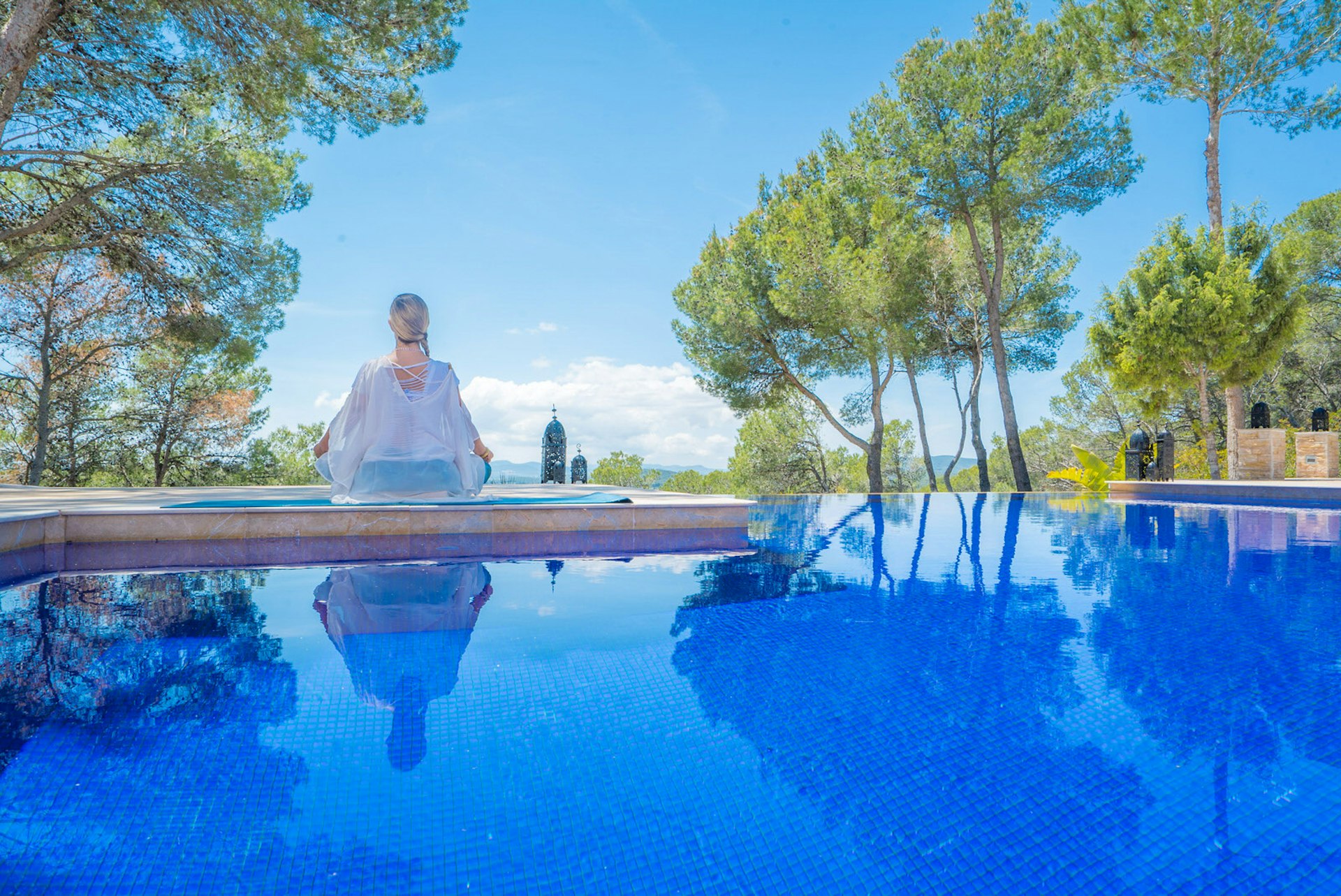 Ibiza spa breaks - a blonde person sits cross-legged in meditation with their back to the camera, next to a pristine blue pool