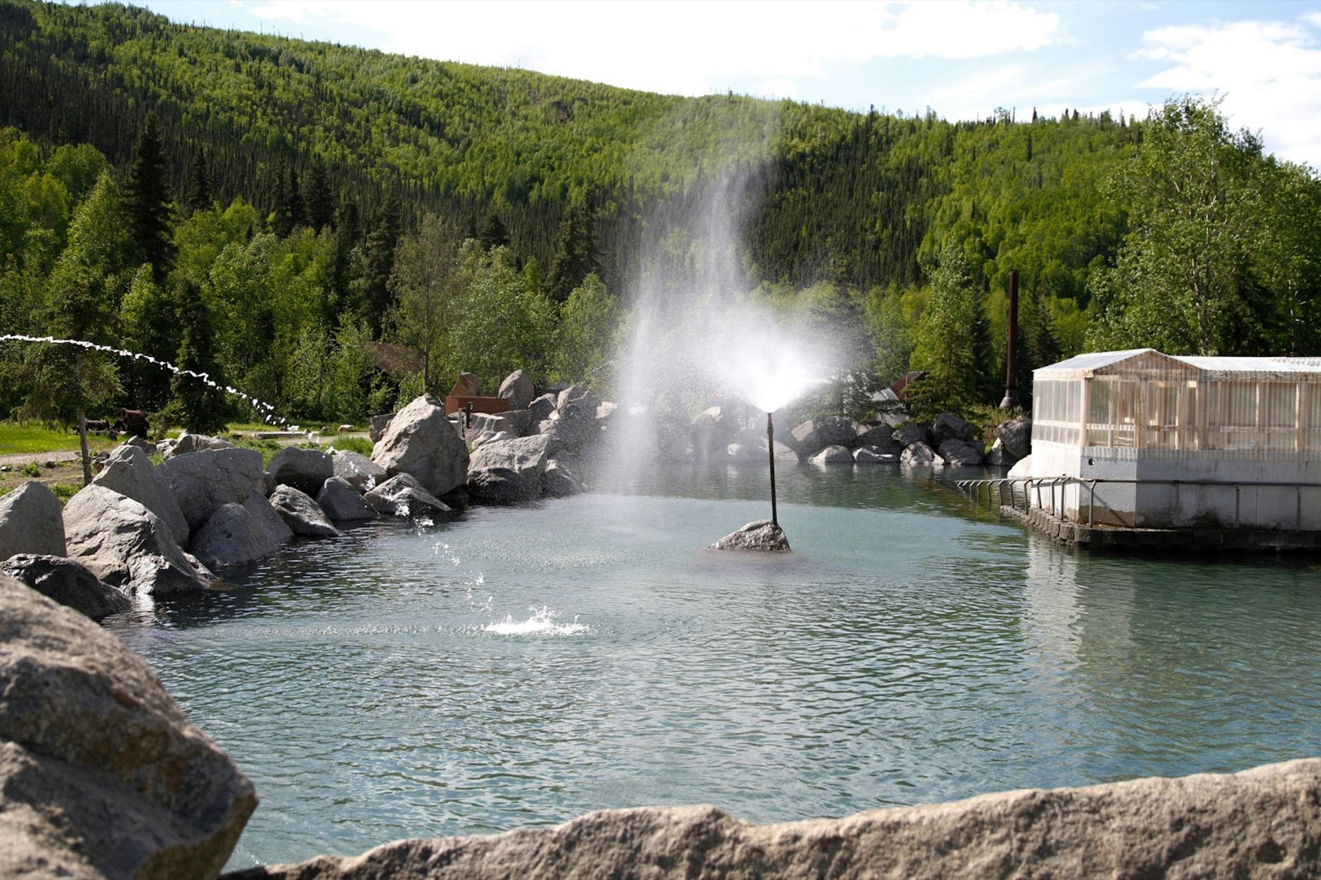 Two fountains spurt water in the late at Chena hot springs, with boulders and a small building around the perimeter