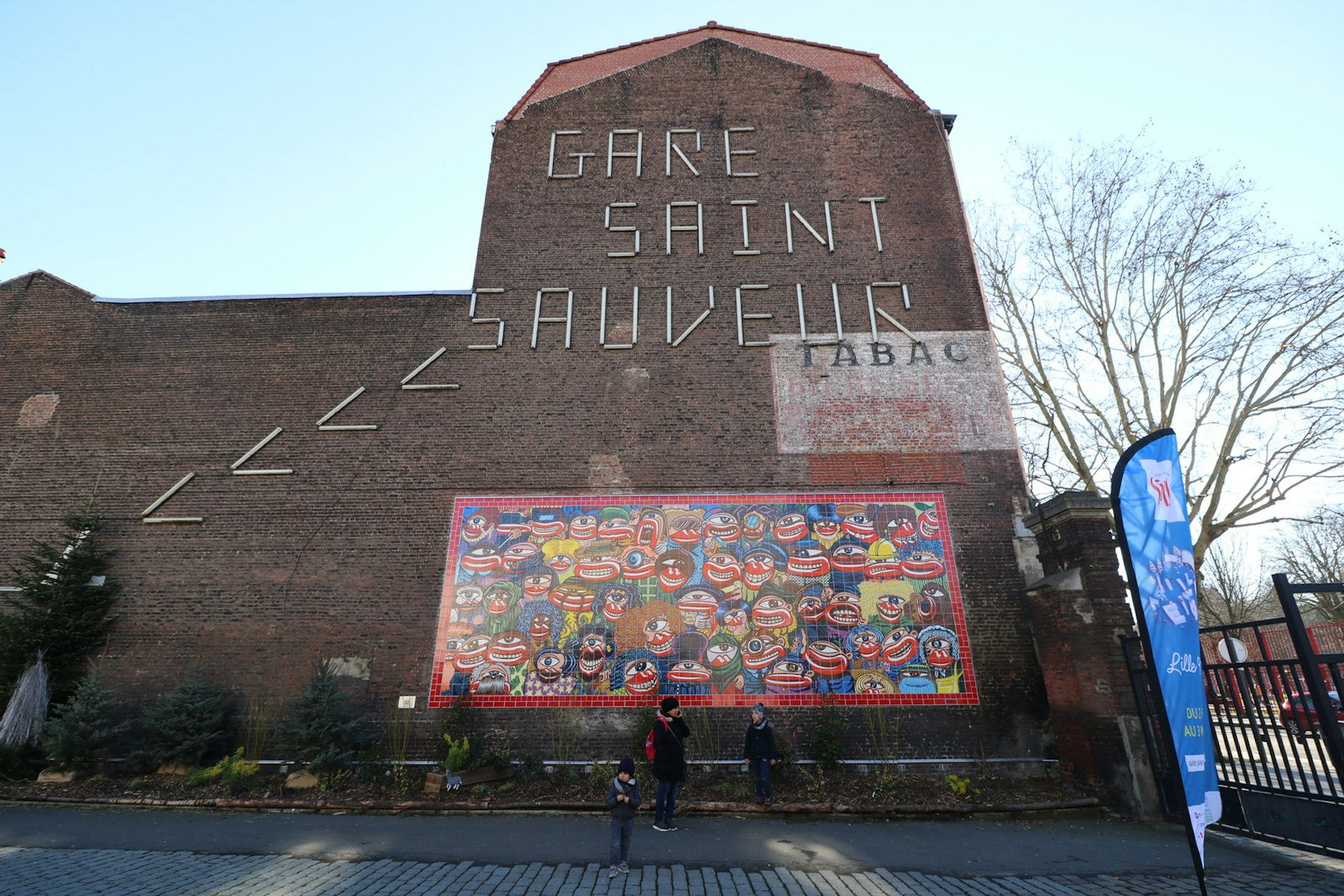 The exterior of a large red-brick building labelled Gare Saint Sauveur, with a colourful mural of eyes and teeth