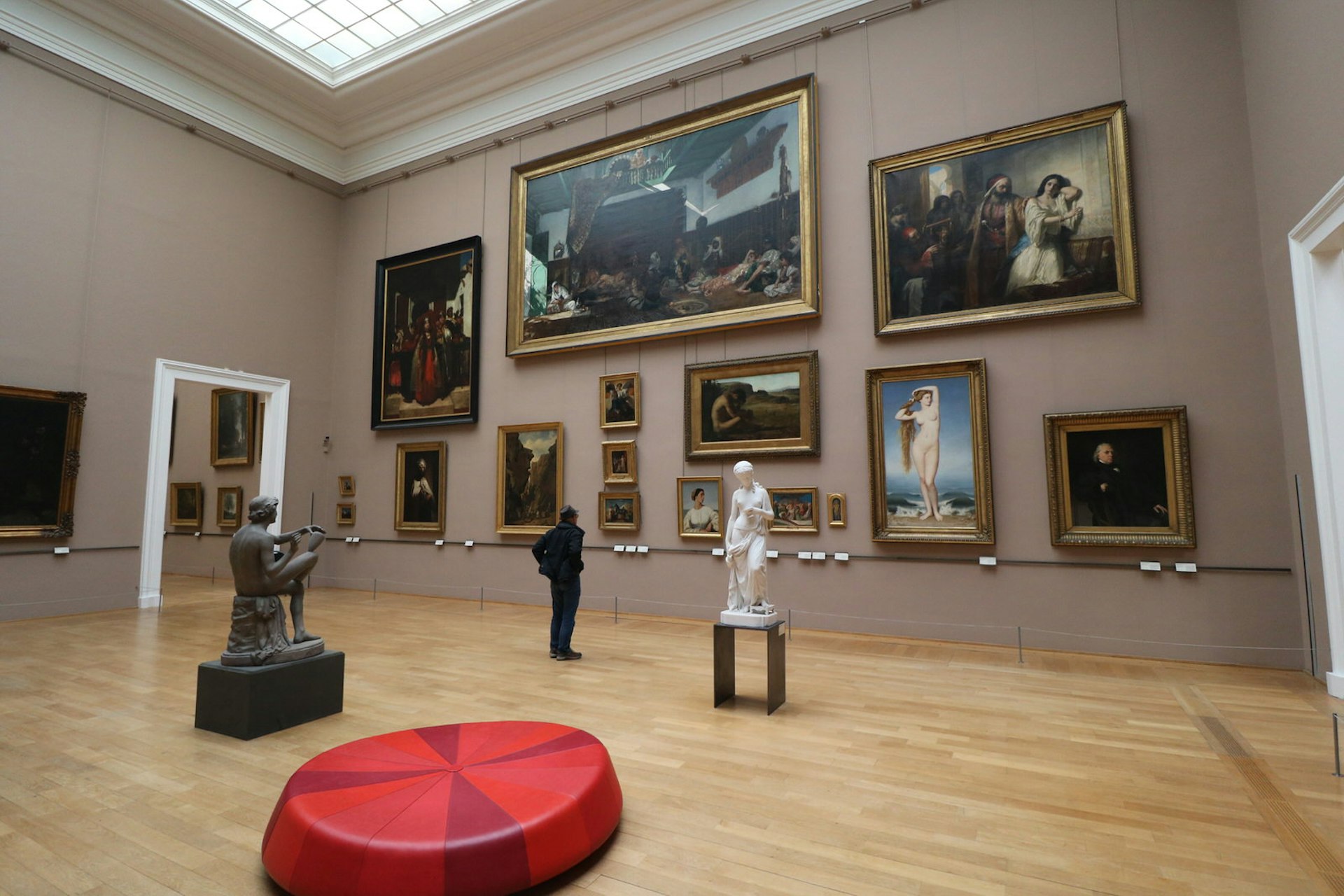 A spectator, flanked by two large sculptures of people, admires the large paintings covered the wall in the Palais des Beaux Arts, Lille