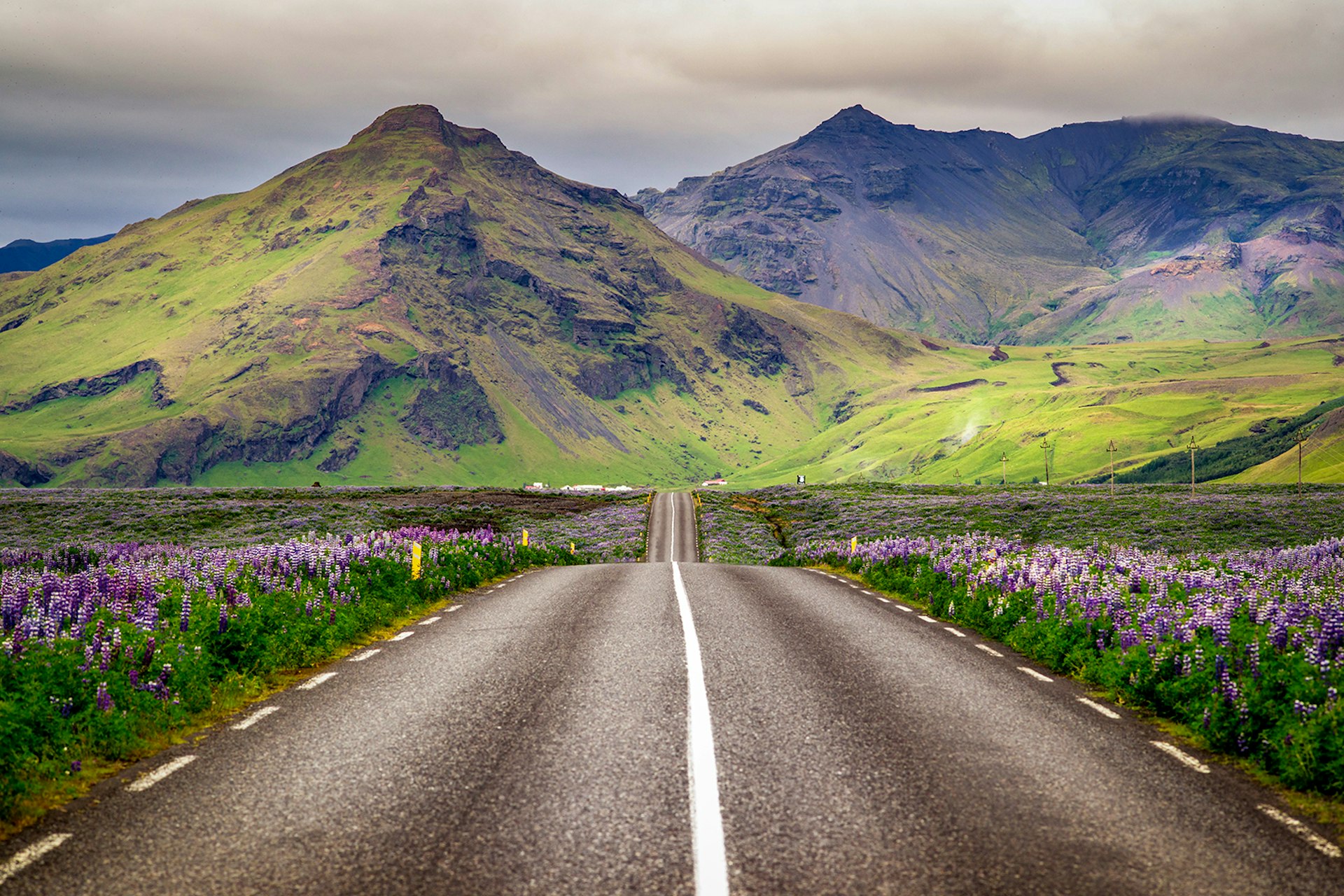 A two-lane road stretches straight ahead towards green, grey and purple hued mountains. Iceland.