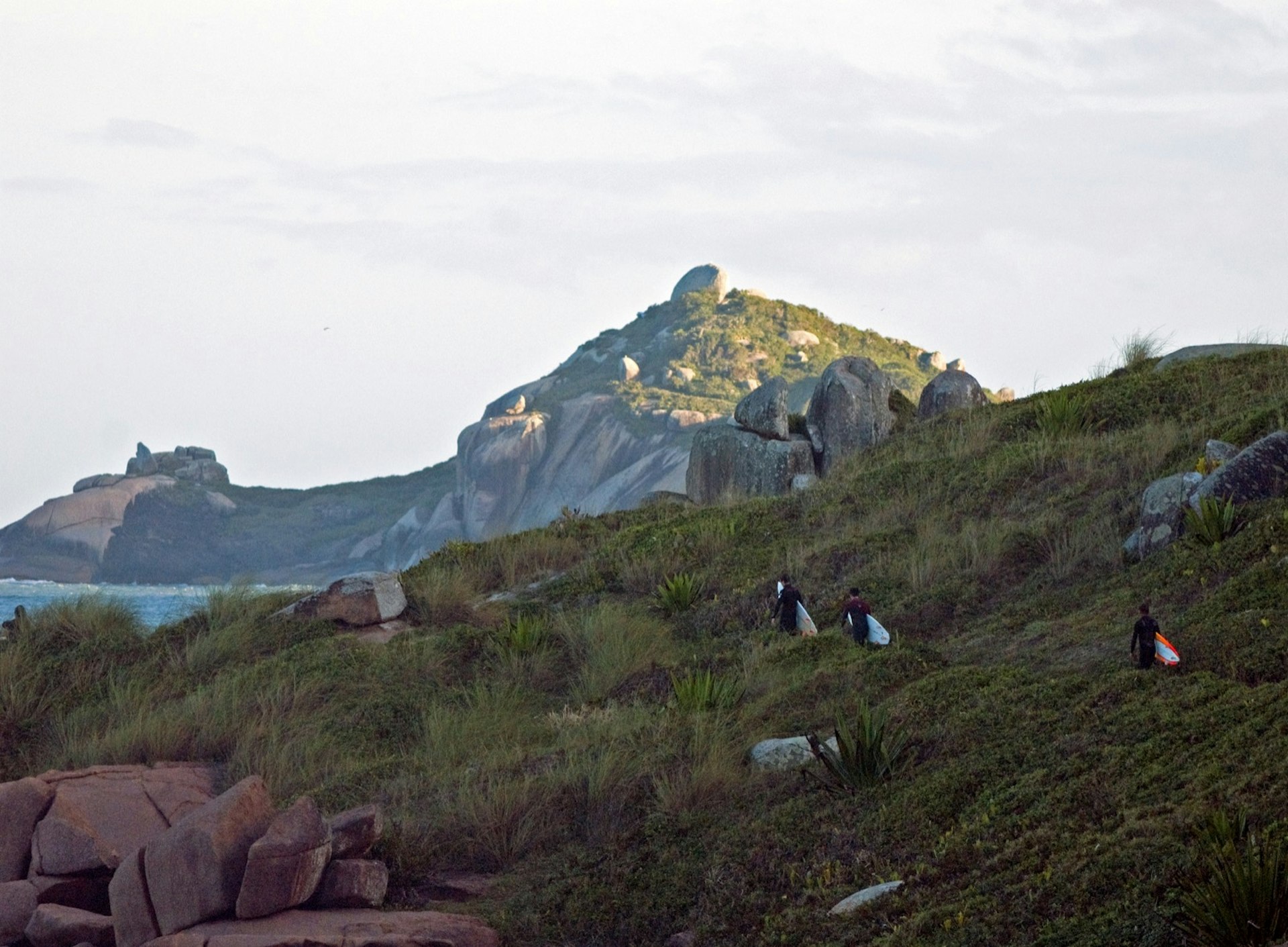 A line of three men carry surf boards up a rocky slope on their way to a spot to surf