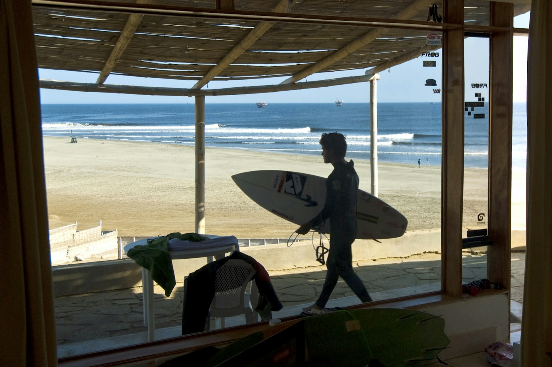A person carries a surfboard across a shady porch at a cabin or cabana situated right on a beach