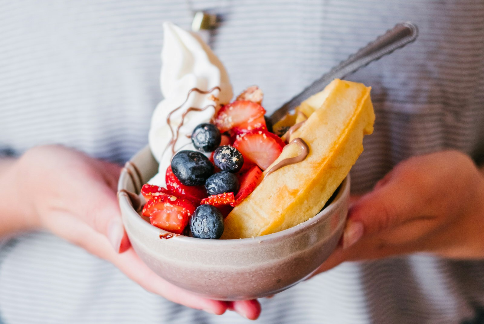A bowl heaped with strawberries, blueberries, vegan cream and a waffle; the bowl cupped by a pair of hands