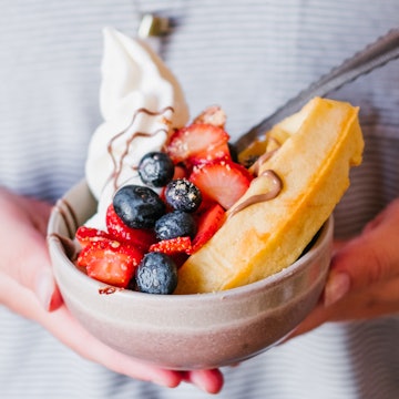 A bowl heaped with strawberries, blueberries, vegan cream and a waffle; the bowl cupped by a pair of hands
