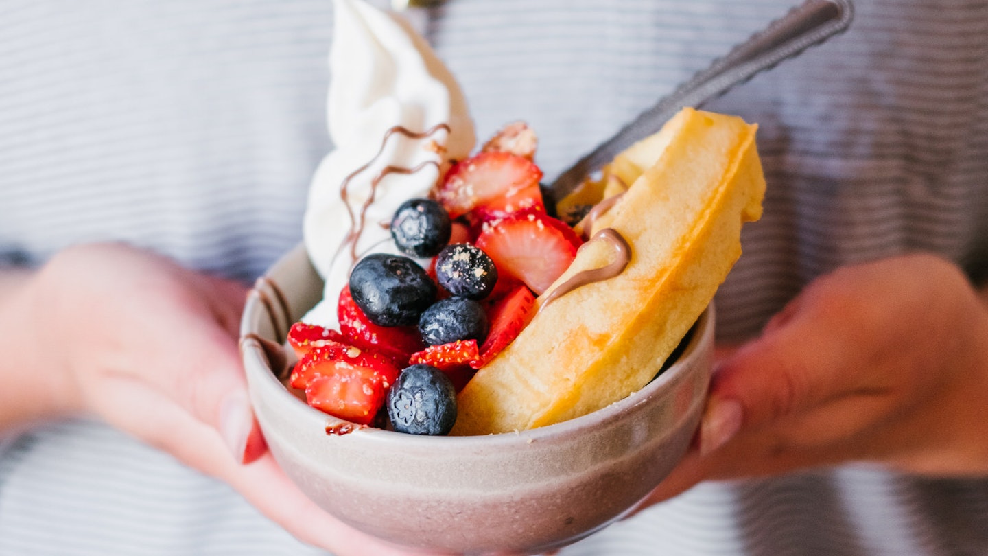 A bowl heaped with strawberries, blueberries, vegan cream and a waffle; the bowl cupped by a pair of hands