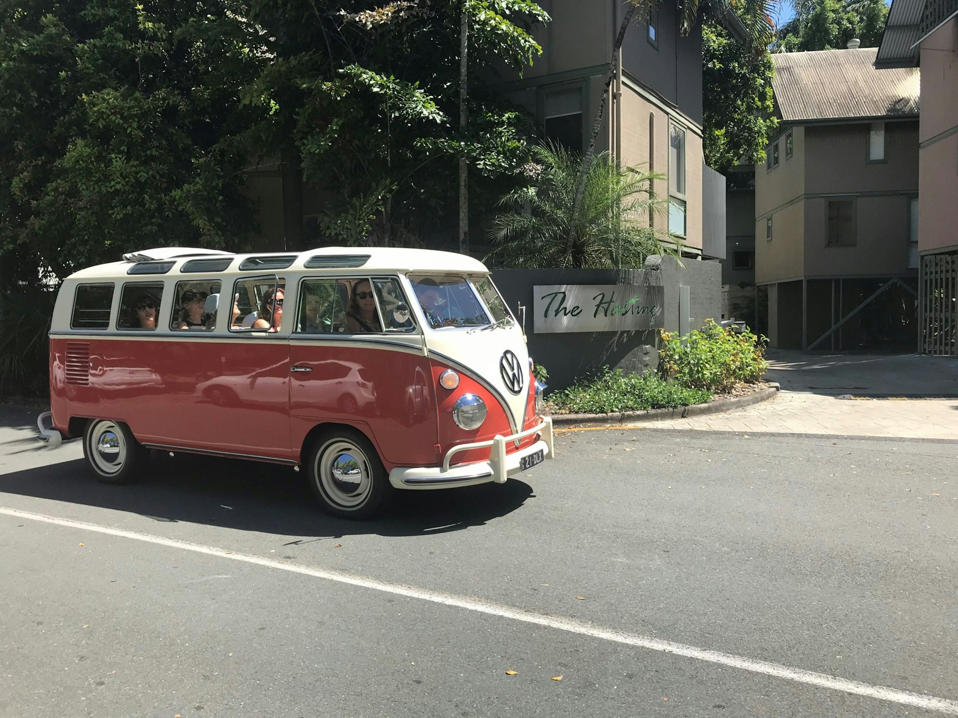 A retro VW Kombi van, with bright red bottom and white top (which converge to a point at the front bumber), drives along a tree-lined street in Noosa; there are numerous individual windows along the van's side and on the edge of the roof, and the middle of the roof is retractable, with the fabric piled near the rear
