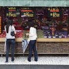 Two people stand in front of a wide range of fruit, displayed below large price signs