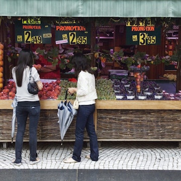 Two people stand in front of a wide range of fruit, displayed below large price signs