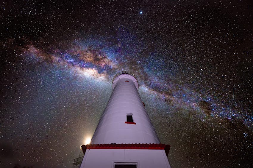 A purplish glow lights the exterior of the white Lady Elliot Island lighthouse, which rises and narrows as it climbs skyward to the heavens above; the black sky is cut from left to right by blue, gold and purple hues of the Milky Way