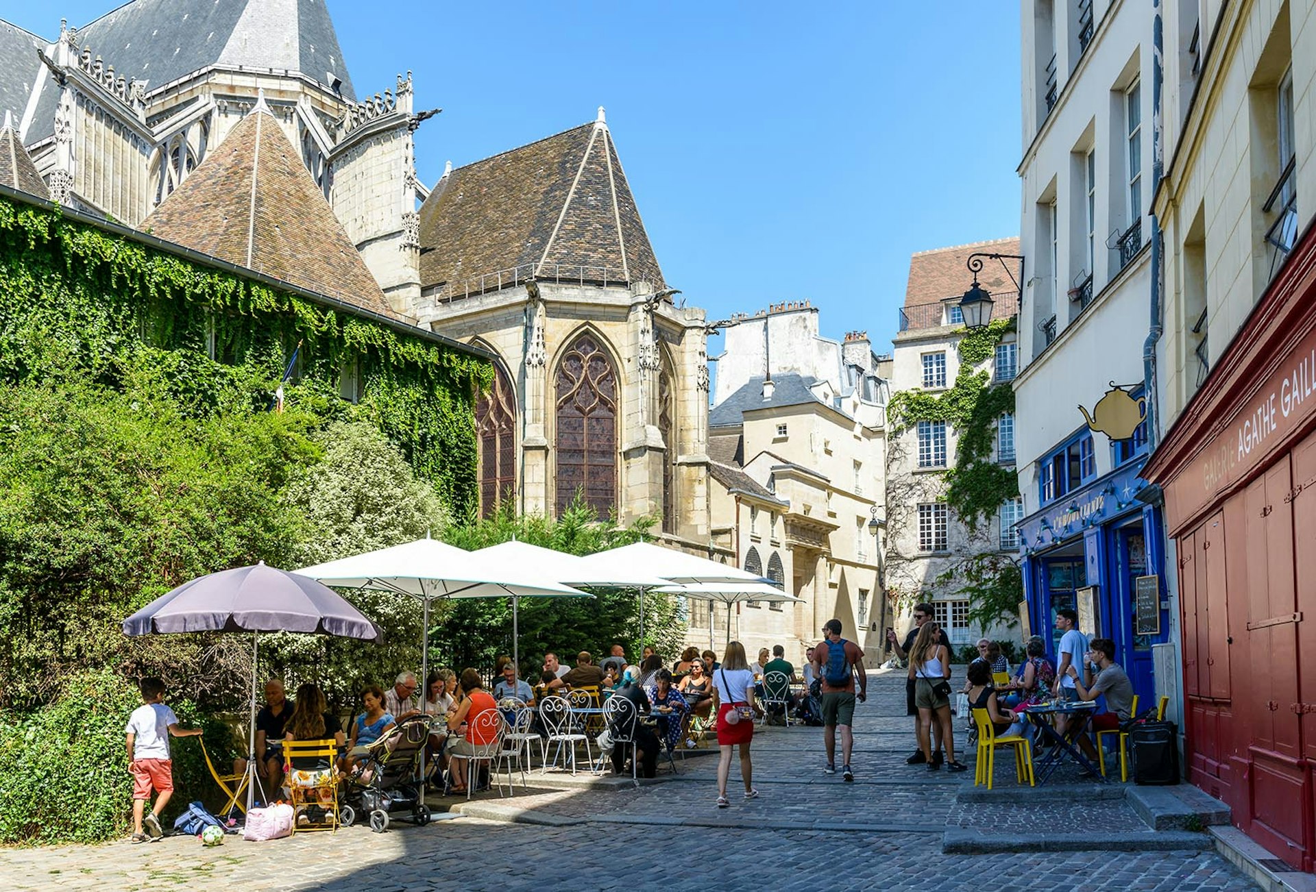 A small pedestrian cobbled street in the old district of Le Marais in Paris, France, behind Saint-Gervais church, with people having lunch on the shady terrace of a restaurant by a sunny summer day