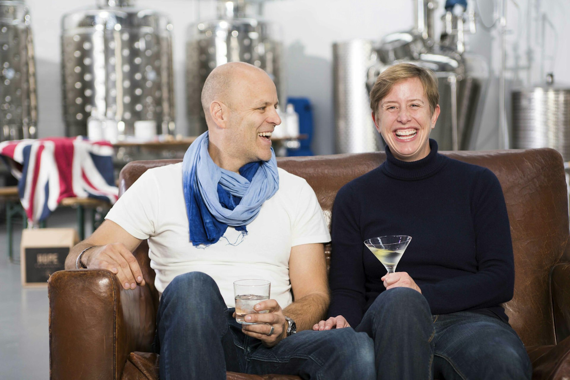 Cape Town gin - Leigh Lisk, a middle-aged man wearing a white t-shirt and jeans (and a big blue scarf around his neck, looks to his left to Lucy Beard, a middle-aged woman with short hair, who is in a black turtle-neck and jeans; she is bursting with laughter, while he chuckles - both of them are sitting on an aged brown leather sofa and are holding glasses
