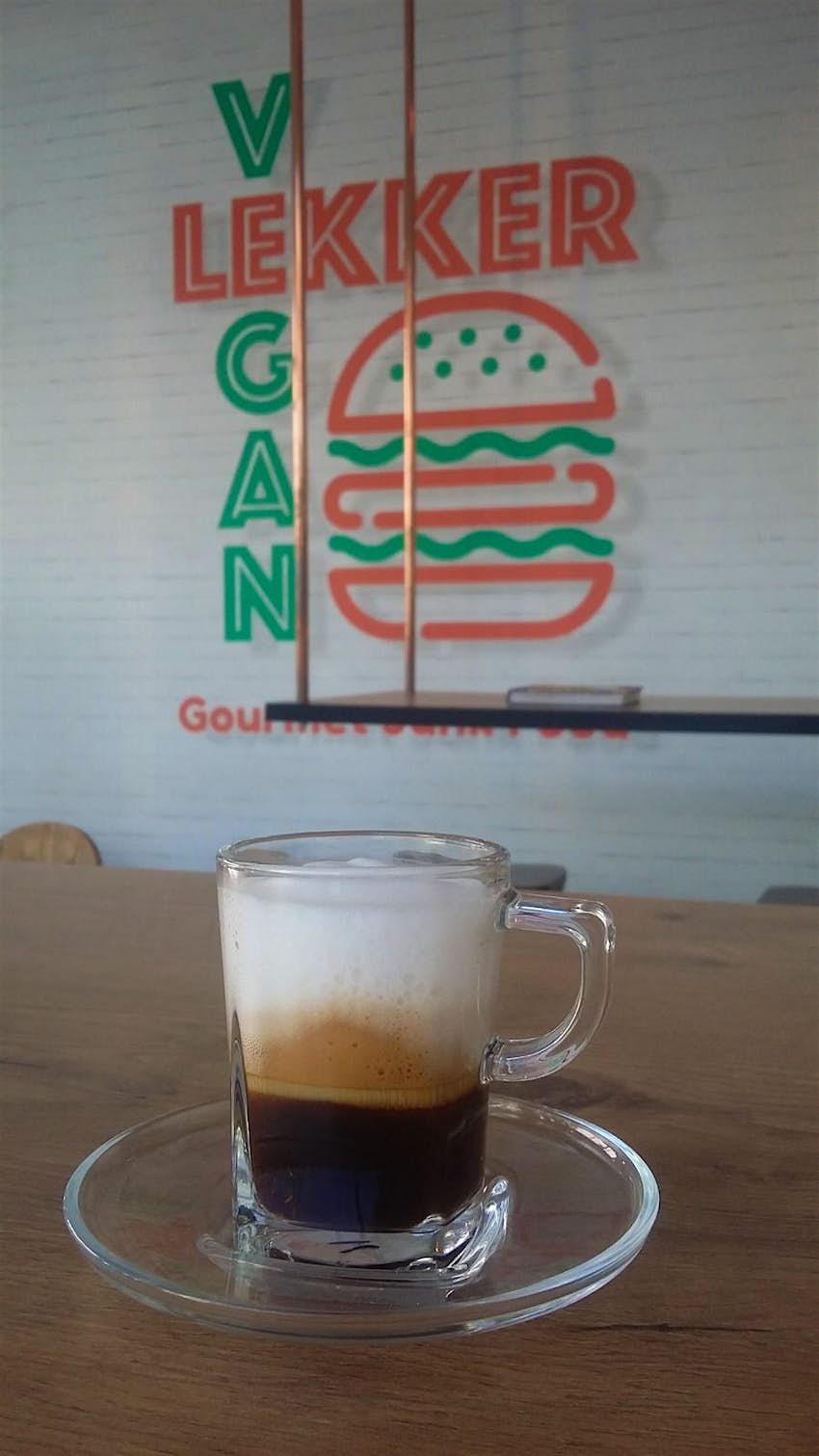 A cappuccino in a clear glass, with its frothy layers on display, sits on a wooden table with the Lekker Vegan logo (a stylised burger with VEGAN written alongside it) in the background; it's one of the best vegan restaurants in Cape Town