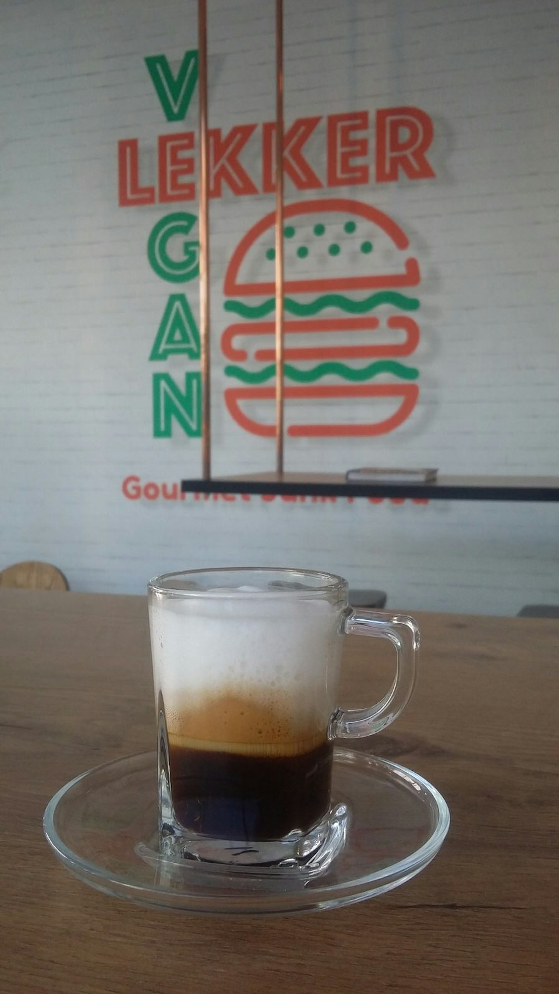 A cappuccino in a clear glass, with its frothy layers on display, sits on a wooden table with the Lekker Vegan logo (a stylised burger with VEGAN written alongside it) in the background; it's one of the best vegan restaurants in Cape Town