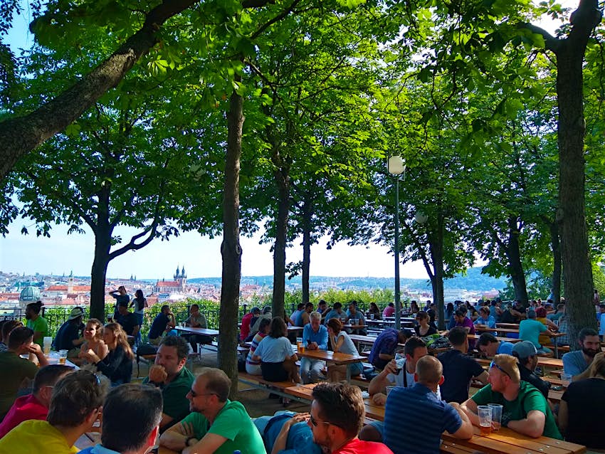 Rows of tables full of beer-drinking people are arranged under a shady canopy of trees on a bluff that overlooks the city of Prague
