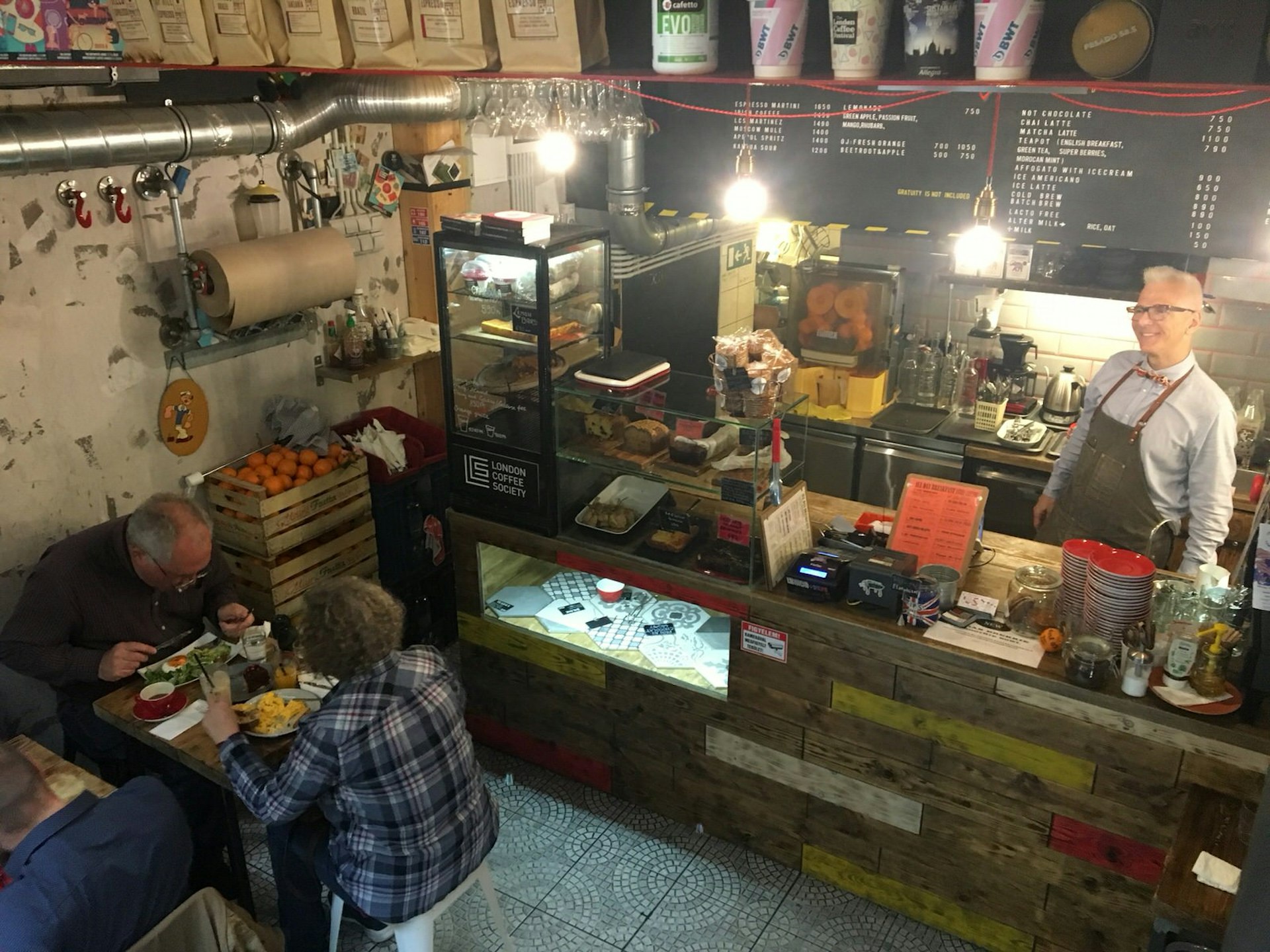 Looking down at the front counter of London Coffee Society from a mezzanine, a man behind the counter is smiling broadly while customers dine on their Budapest brunch