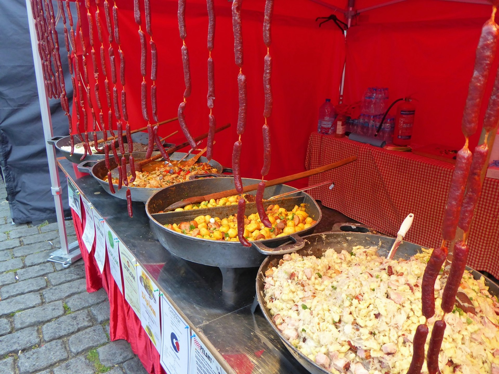 A market stall features four huge pans of food, with long-handled wooden spoons to stir them, and long strings of sausages hung vertically in the front