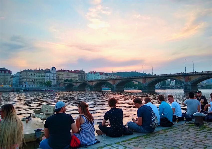 A row of young people sit on the concrete riverbank, looking at an old-world bridge across the river in Prague