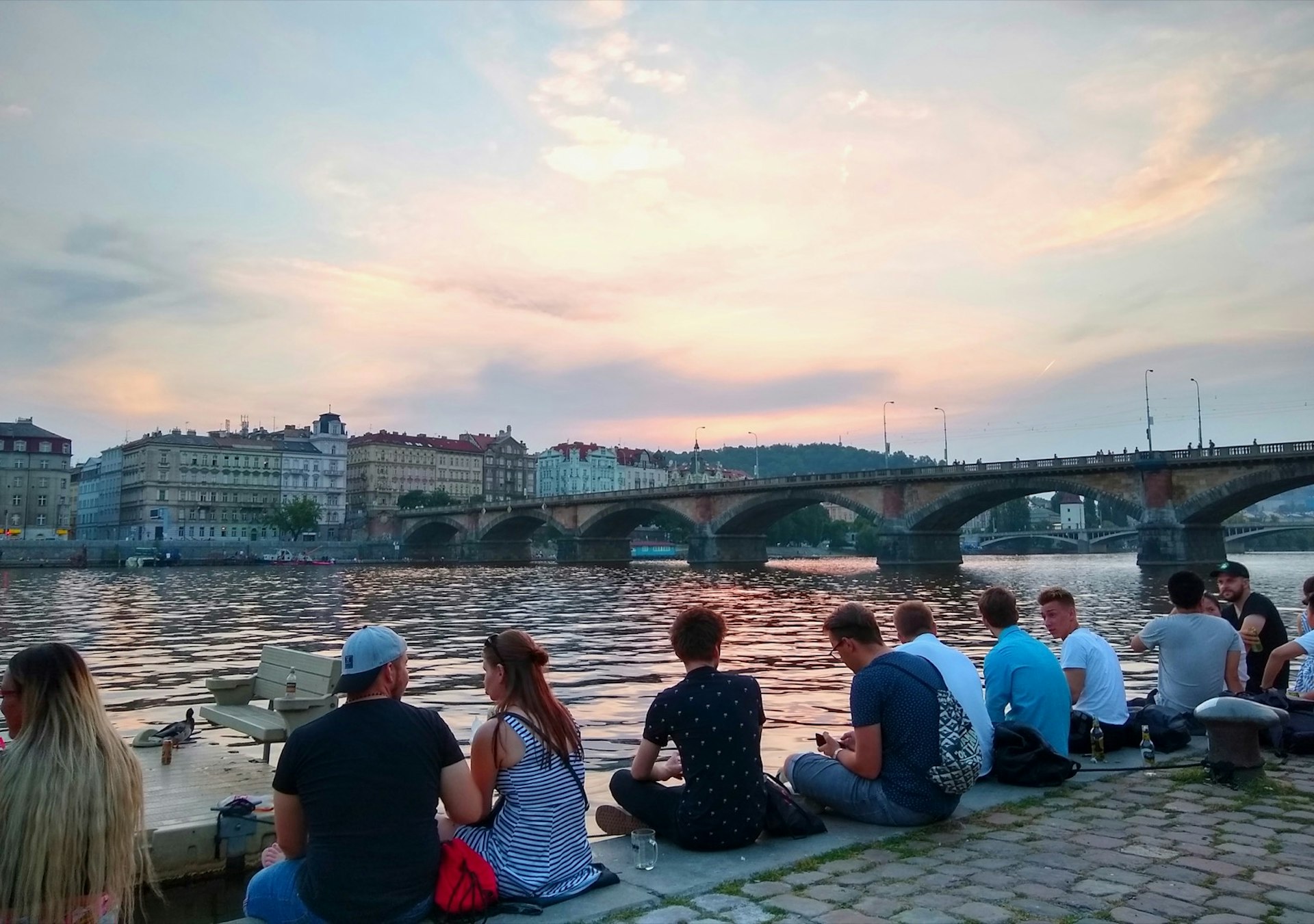 A row of young people sit on the concrete riverbank, looking at an old-world bridge across the river in Prague