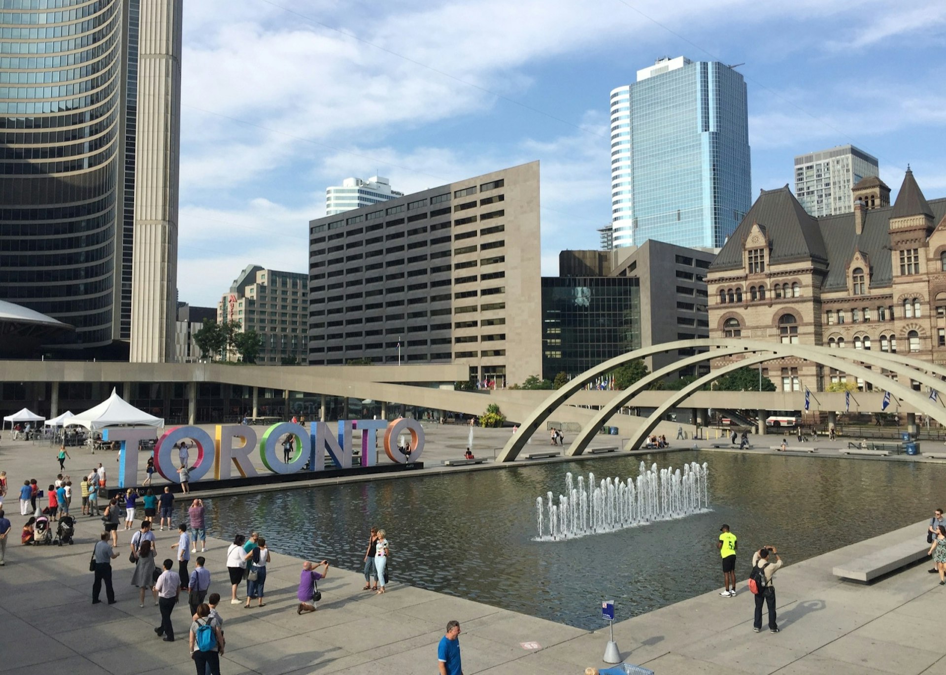 People interact around a large fountain, with giant rainbow colored letters spelling out Toronto nearby