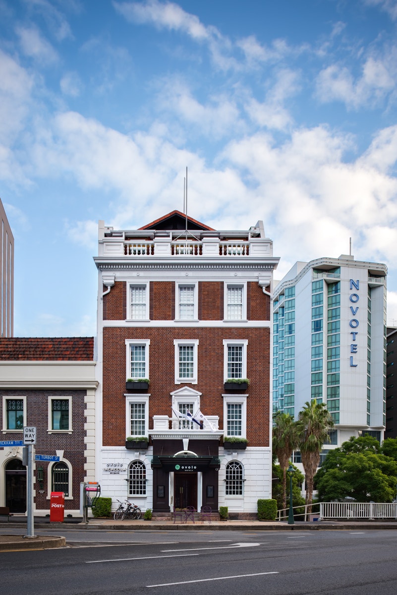 A narrow, but tall, colonial red-brick building (with white-trimmed wood-framed windows and bushy window boxes) stands next to the street, with a large glass high-rise hotel in the background; the ground floor facade of the historic building is white-stone with arched windows and grand entrance