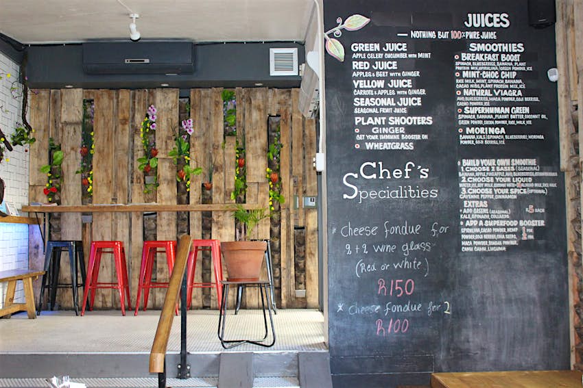 A large chalkboard runs down the right side of the image, with lists of juices, smoothies and chef's specialities; to the left is a wall made of roughly hewn vertical wood planks that are separated with potted plants between them, a counter sits midway up the wall with colourful metal stools; Plant is known as one best vegan restaurants in Cape Town
