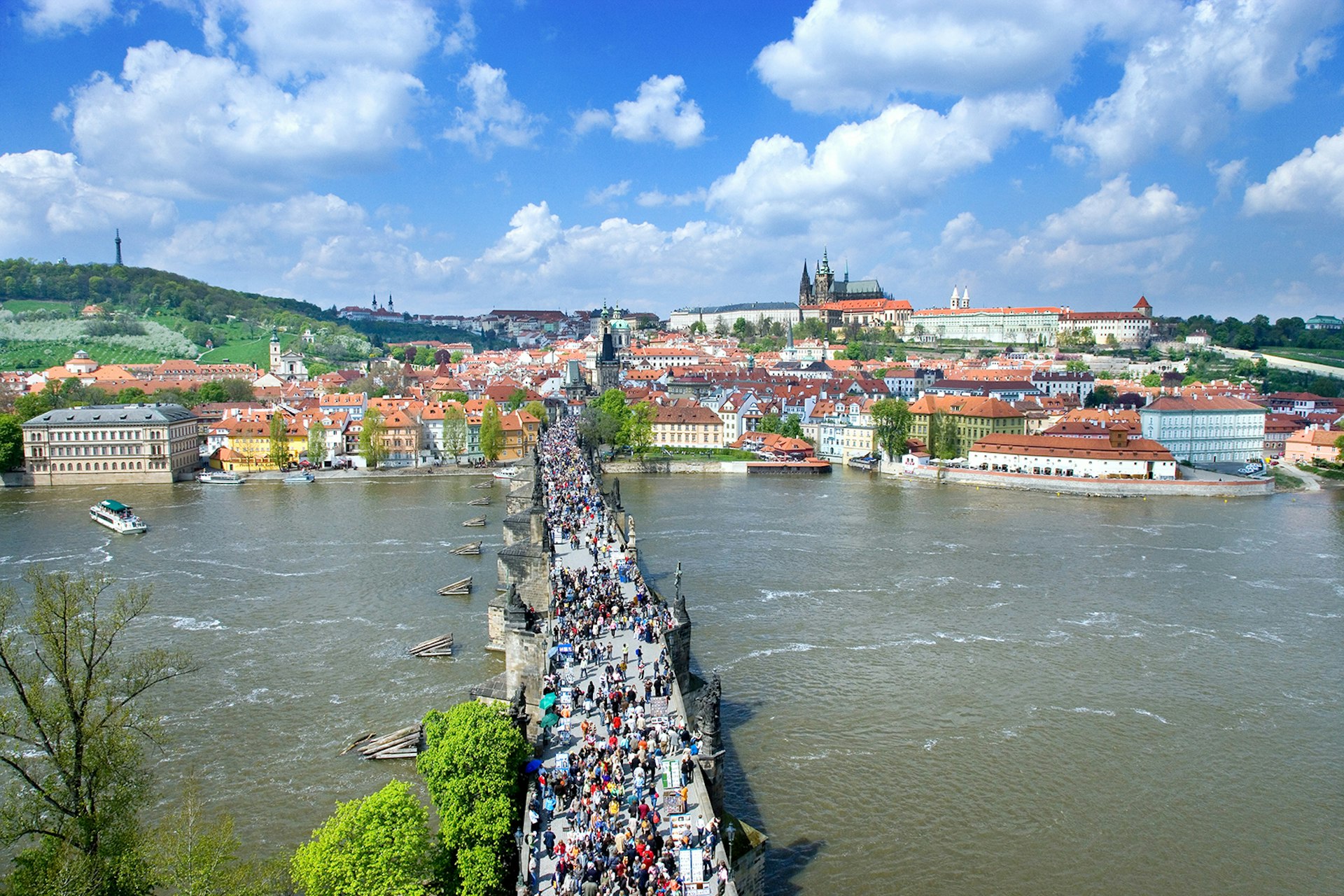 An aerial view of Charles Bridge and people crossing Prague's Vltava River with the castle in the background