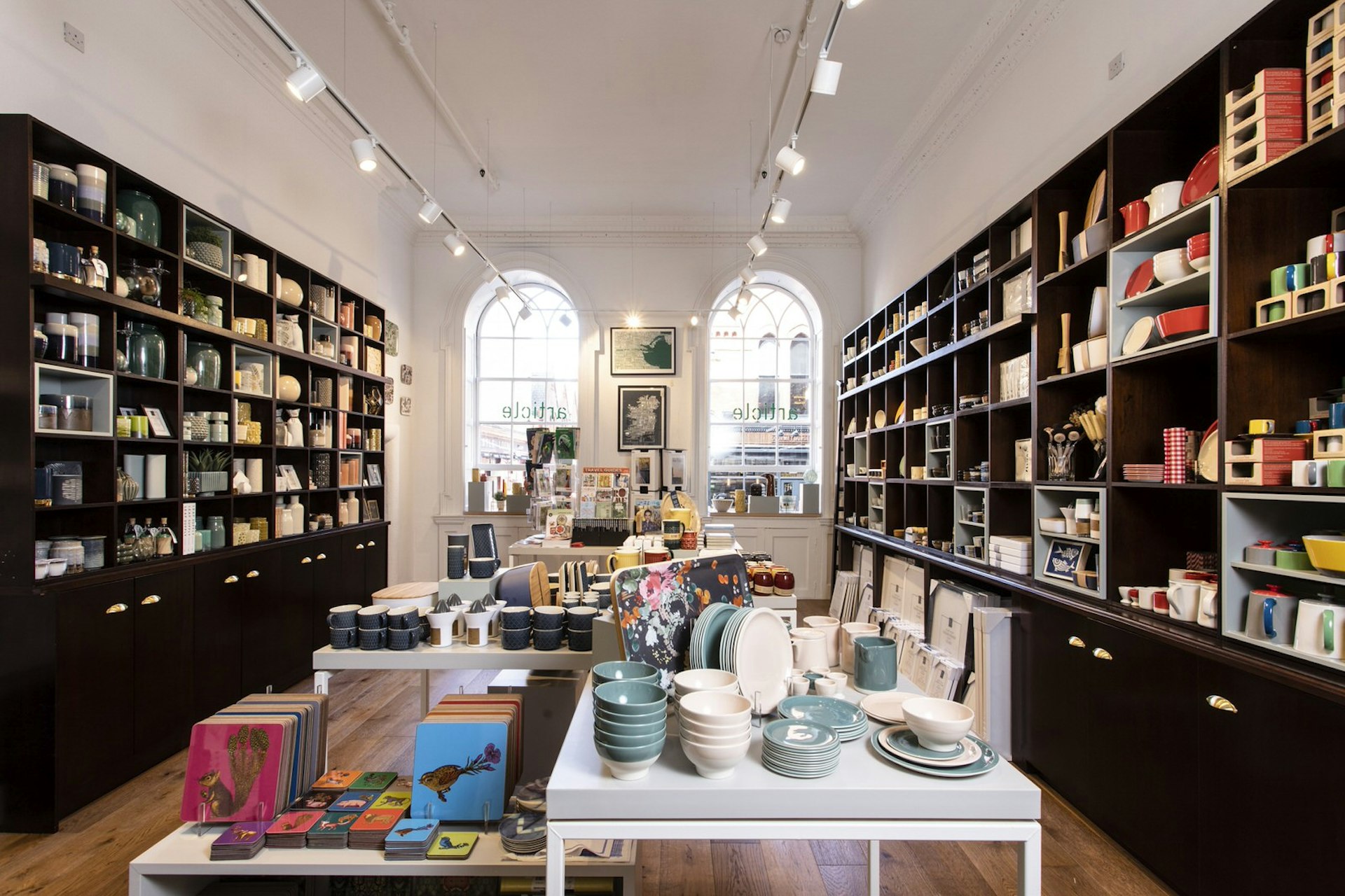 Dublin independent shops - the interior of Article. Tall dark shelves line the white walls stocked with colourful bowls, cups, jugs and homewares. In the foreground there is a white table with notebooks and more bowls. There are large arched windows on the back wall