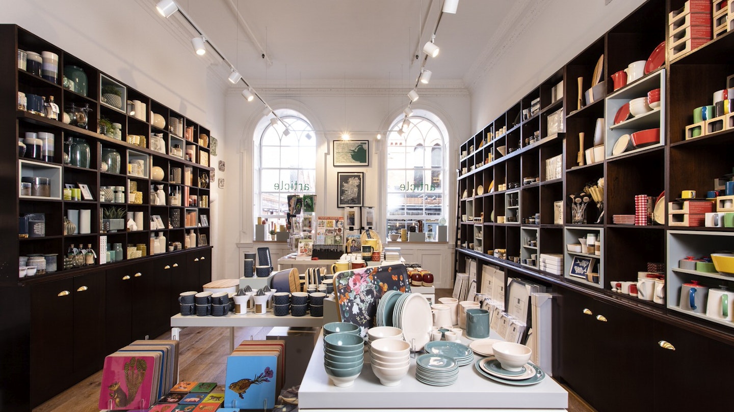 Dublin independent shops - the interior of Article. Tall dark shelves line the white walls stocked with colourful bowls, cups, jugs and homewares. In the foreground there is a white table with notebooks and more bowls. There are large arched windows on the back wall
