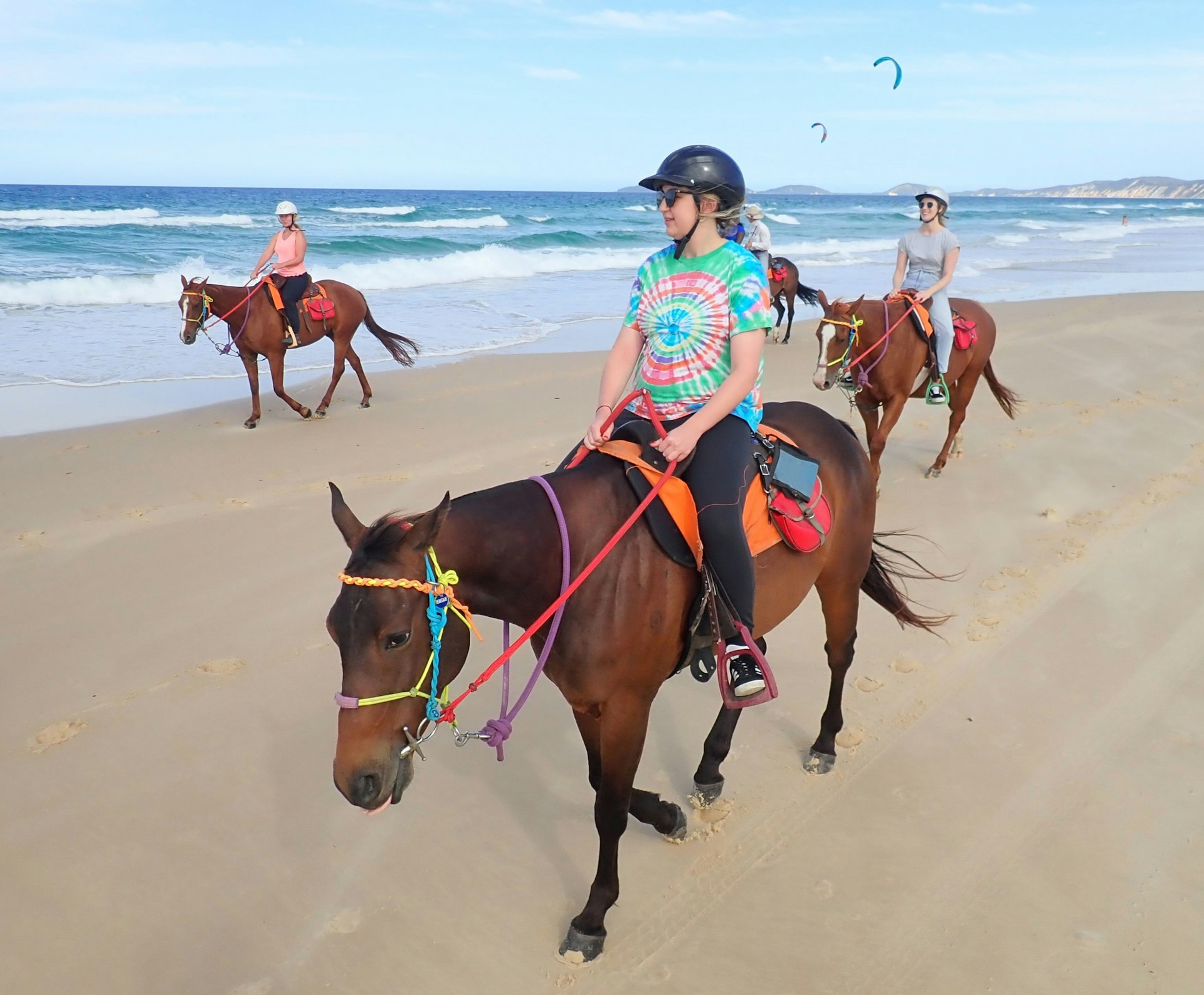 A young woman wearing a tie-died t-shirt, black grousers, helmet and sunglasses rides a chesnut-coloured horse with brighly coloured reigns, which range from purple and orange to red, blue and green; behind her are two other riders on the same section of beach, with crashing waves beyond and two kite-surfers in the distance