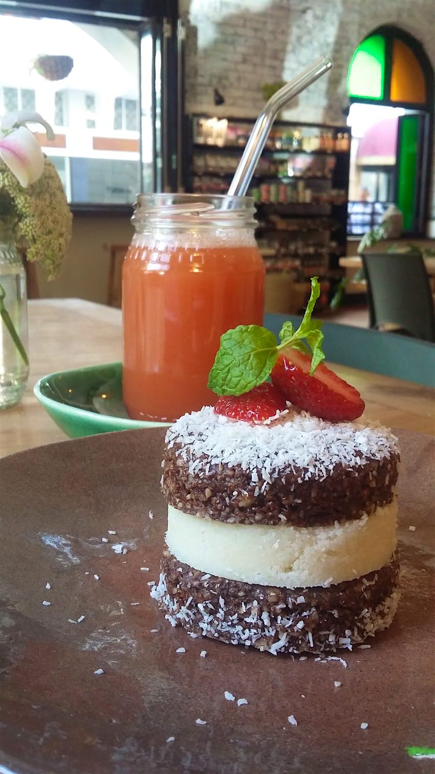 A jam jar full of tomato-looking juice sits behind a three-layered desert; the chocolate top and bottom are separated by a creamy filling; atop the layers is desiccated coconut and two slices of strawberry; with dishes like this, it's easy to see why Raw & Roxy is one of the best vegan restaurants in Cape Town