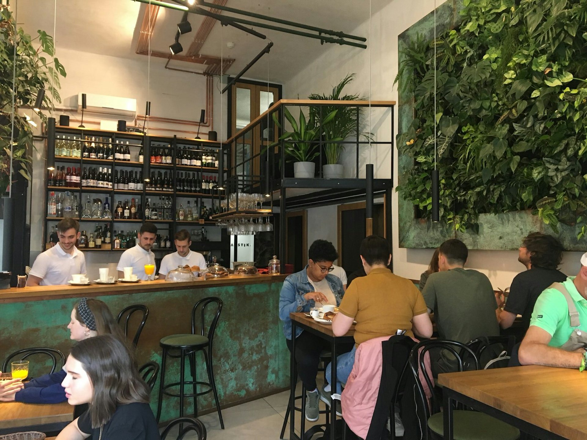 Three white-shirted baristas stand behind a tall counter that is lined with glasses of coffee and juice; behind them is a tall wall of shelves lined with bottles of wine; there is a wall of plants, as well as several shelves with large potted plants; in the foreground are diners enjoying their Budapest brunch while seated at tables