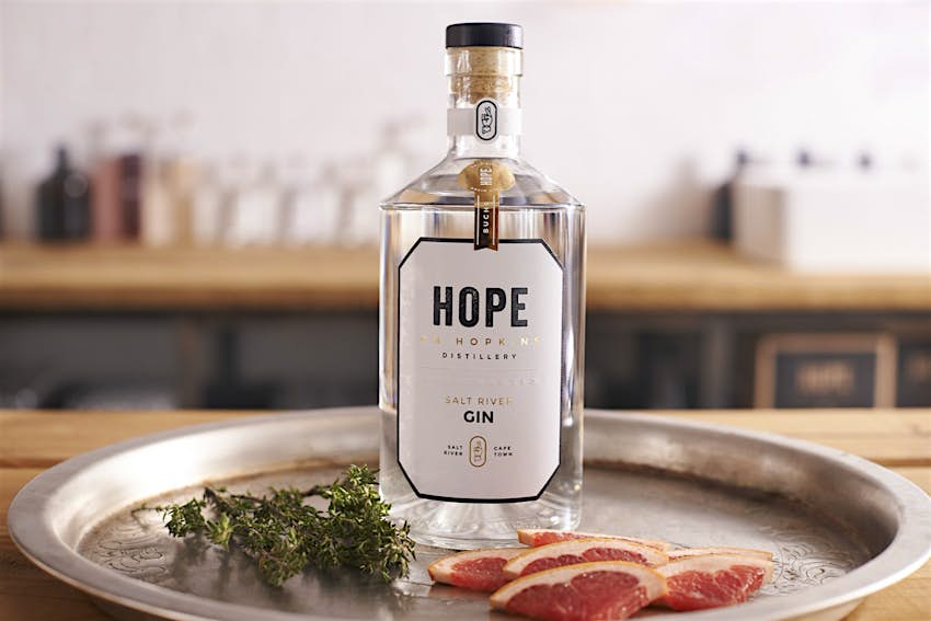 Cape Town gin - A clear bottle of gin, with a corked top and white label with 'HOPE' emblazoned across it, sits on an aged silver platter with some rosemary and pink grapefruit slices; in the background is a very blurred wooden countertop with with various glass vessels on it