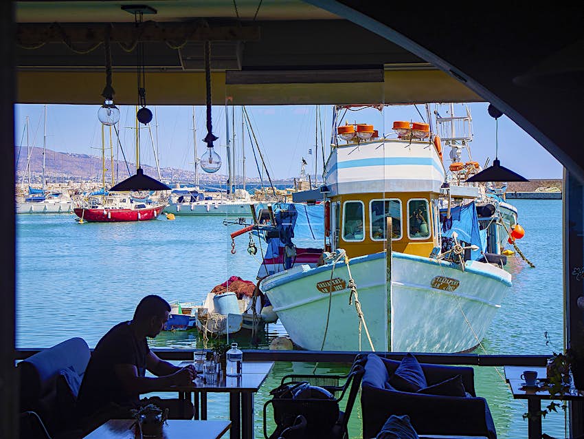 A view from inside a restaurant looking out at a harbor; a man sits at a table in front of a moored fishing boat in Piraeus, Greece.