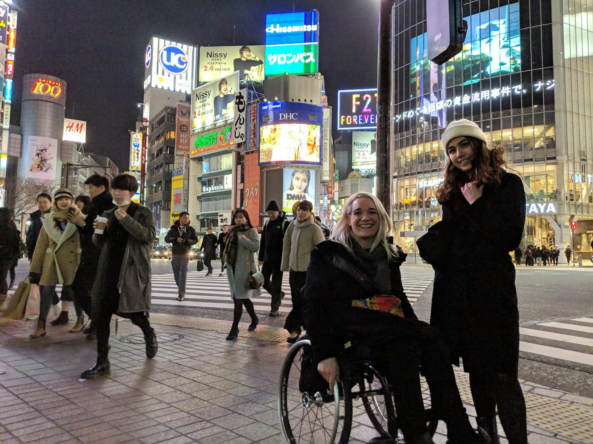Writer Louise Bruton, a wheelchair user, and her friend posing for a photo at Tokyo's Shibuya Crossing; Japanese people are crossing behind them, with the modern buildings and bright lights of Shibuya in the background.