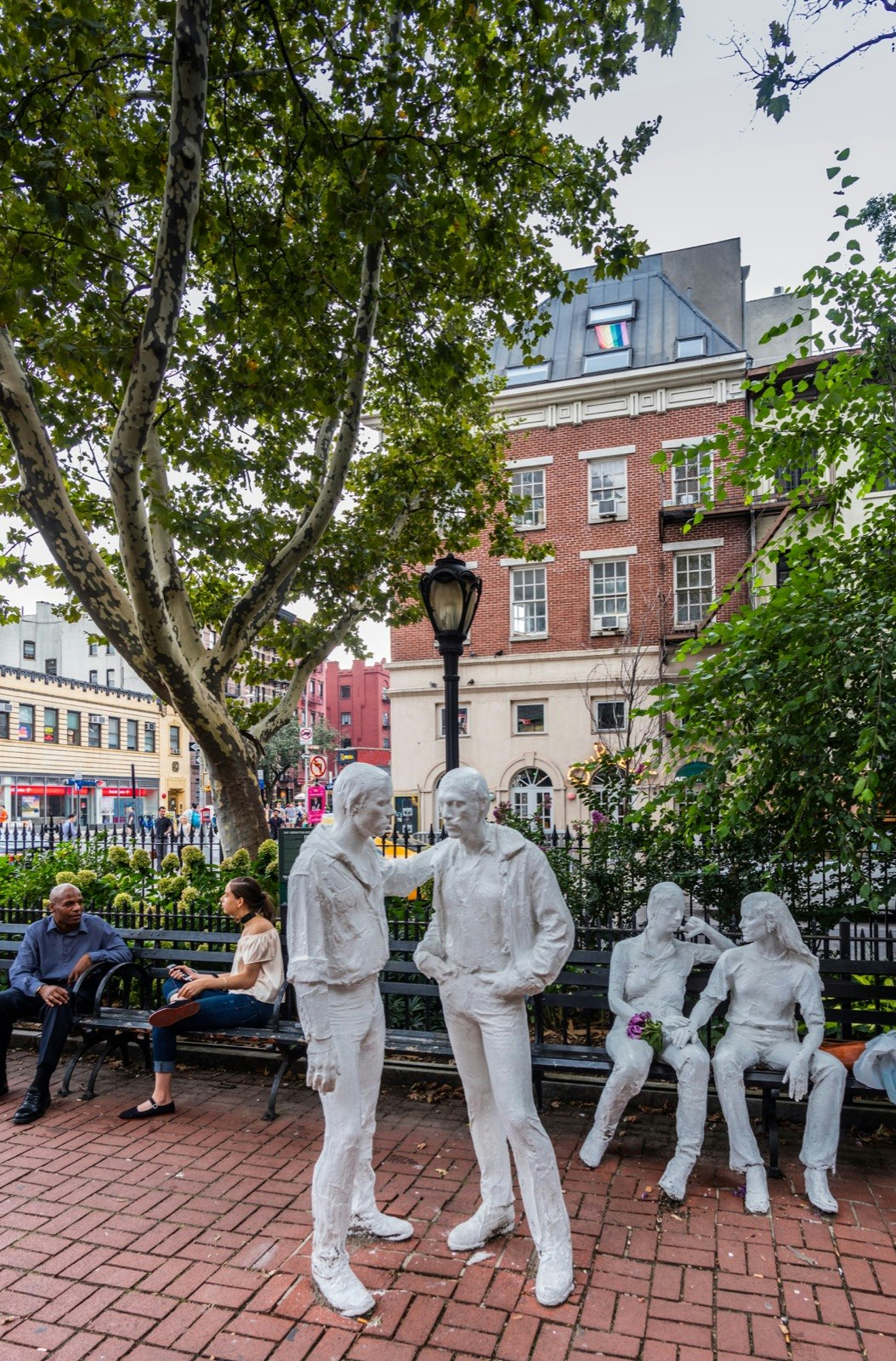 The Stonewall Riot Monument shows four white statues, two standing and two sitting, in a park in New York City. NYC WorldPride will celebrate the 50th anniversary of the Stonewall Uprising. A man and woman sit on a park bench in the background. 