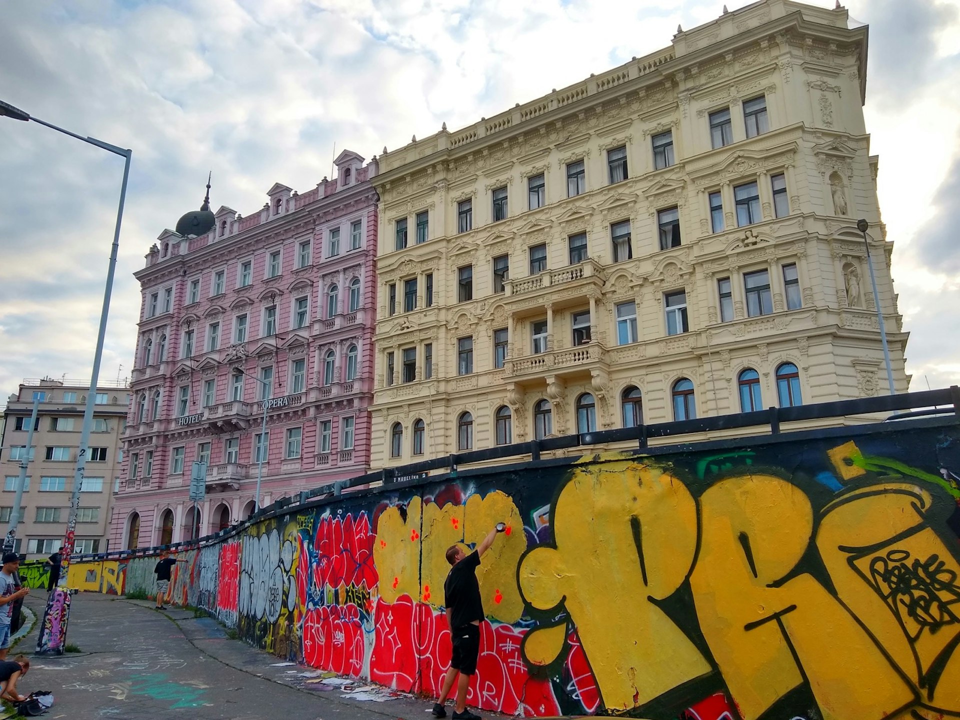 A long row of graffiti marks a wall in Prague, in front of two multi-story apartment buildings, one purple and one cream. 