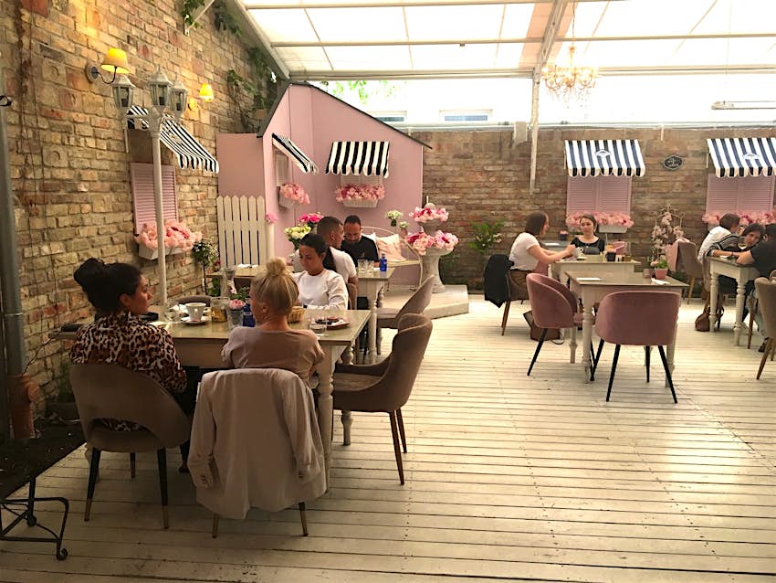 A series of wooden tables with pink chairs, most full of patrons, sits on white floorboards; there are brick walls with closed pink shutters, while light cascades in from a transparent ceiling