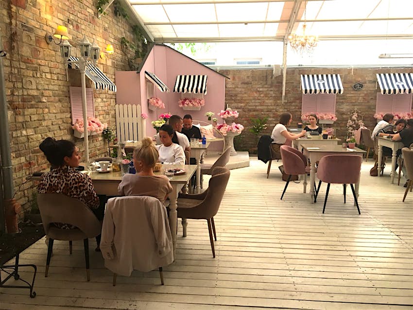 Features - Vintage-Garden--22b9b34cef37 A series of wooden tables with pink chairs, most full of patrons, sits on white floorboards; there are brick walls with closed pink shutters, while light cascades in from a transparent ceiling