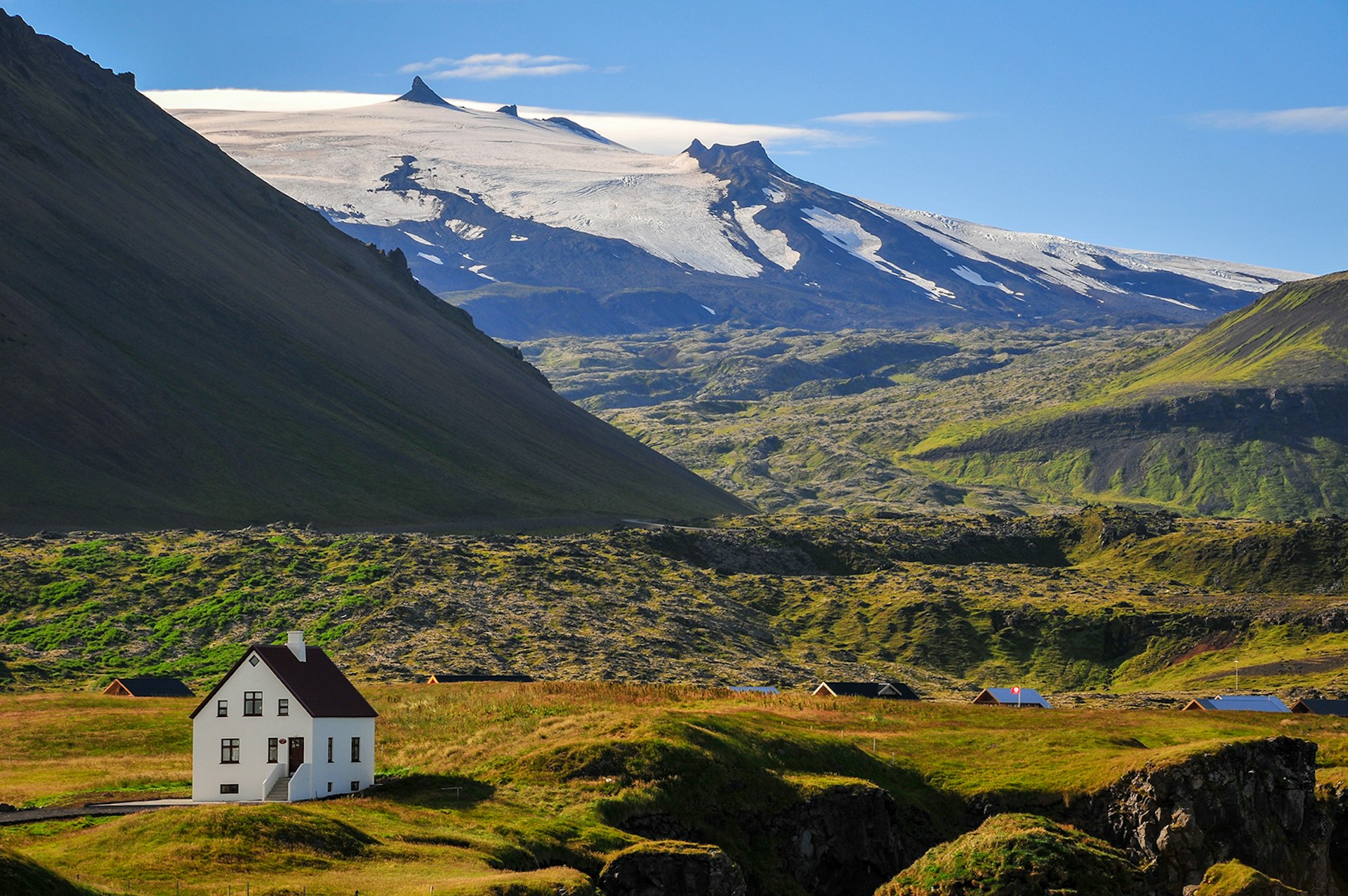 A small white cottage sits on a rolling mossy field with large volcanic mountains and glaciers in the background. West Iceland.