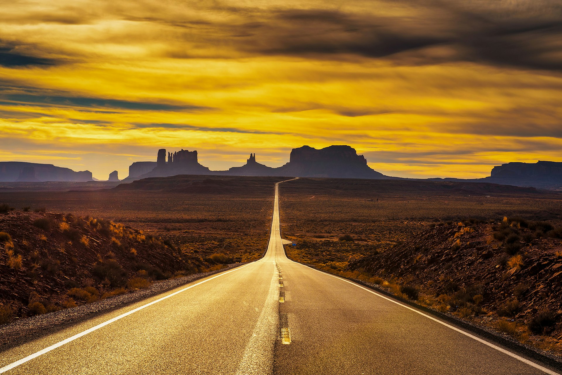 Western US road trip - A highway extends straight towards the rock towers of Monument Valley. The sky turns gold as the sun sets. Western USA.