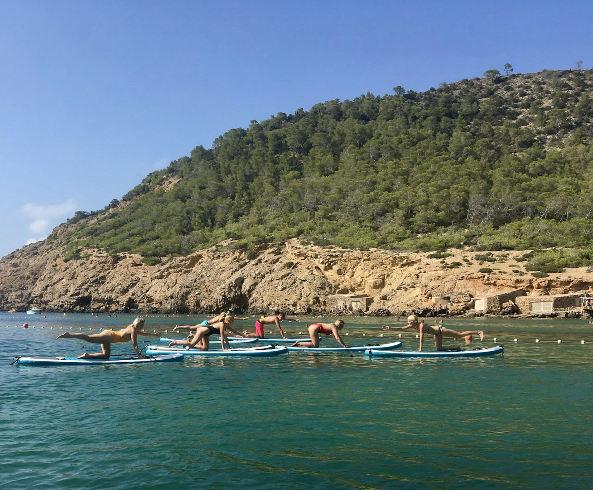 Ibiza spa break - several people practising pilates on stand up paddle boards on a deep blue sea with lush green hills in the background