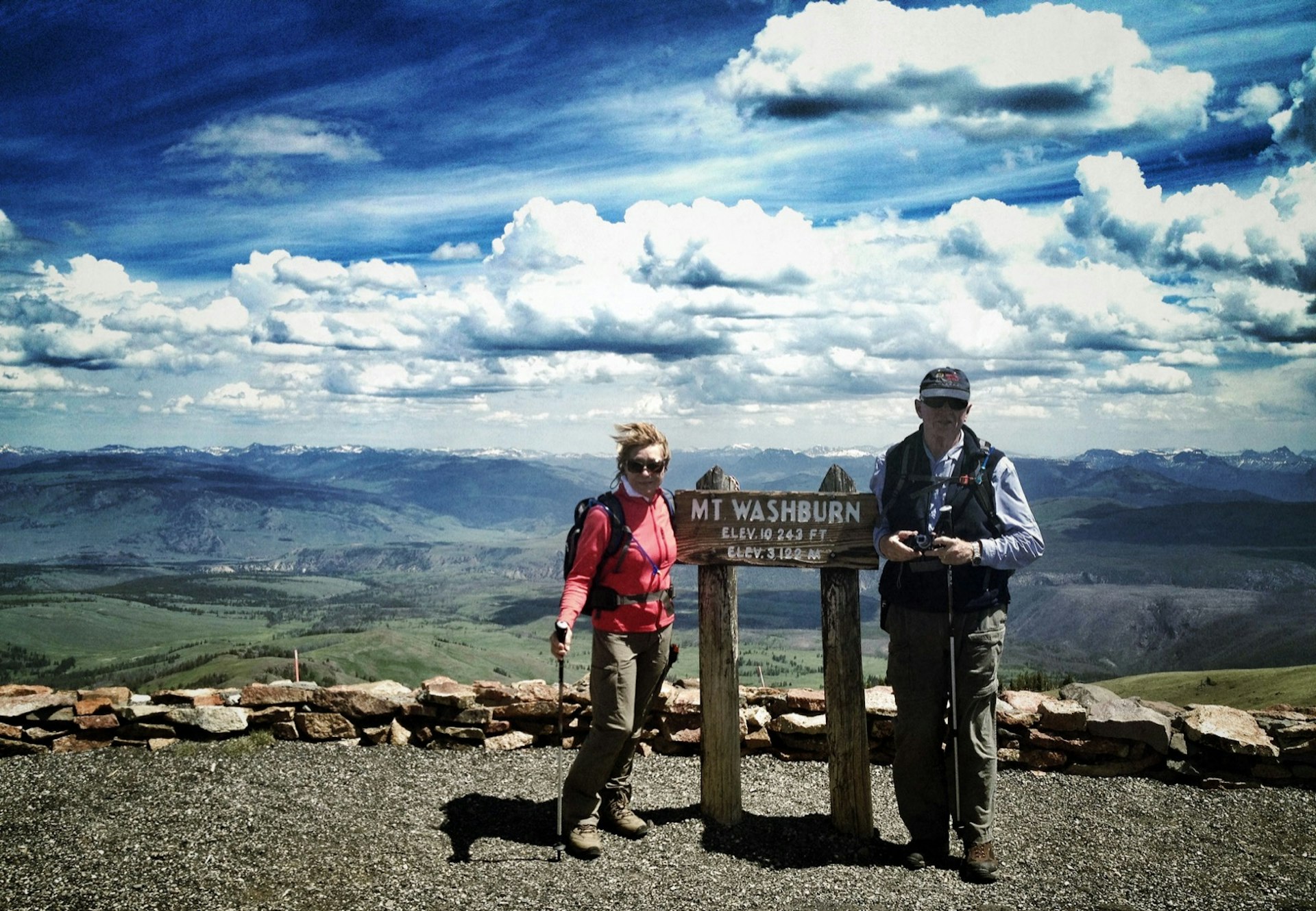 A senior couple stands by a wooden sign at the summit of Mount Washburn in Yellowstone National Park